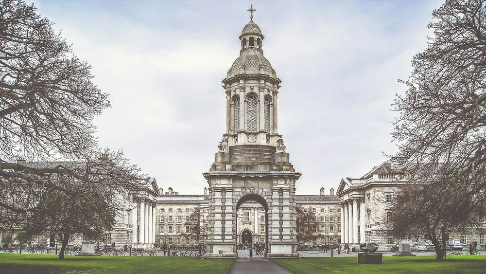 Living In Dublin 2024: The Complete Expat Guide 6 A large tower-like arch near green grassy fields and trees. This is the entrance of the Trinity College in Dublin.