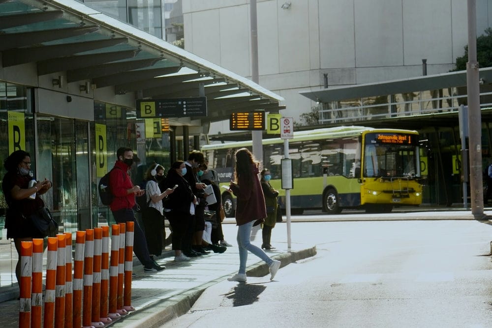 19 Pros And Cons Of Living In Wellington New Zealand 7 A group of people waiting at a bus stop. A yellow bus rushing in their direction can also be seen. Wellington is an eco-friendly city with a strong accent on the natural way of doing things.