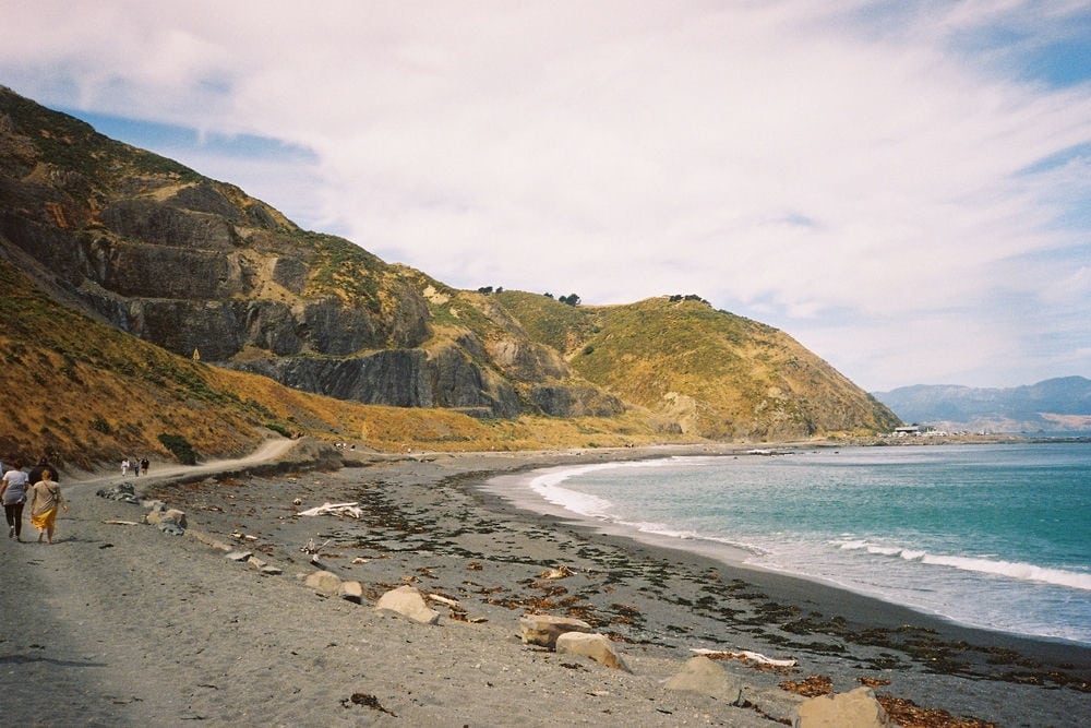 19 Pros And Cons Of Living In Wellington New Zealand 5 Two people walking on a seashore below the mountains under blue skies and clouds. Living in Wellington New Zealand means easy access to beaches.