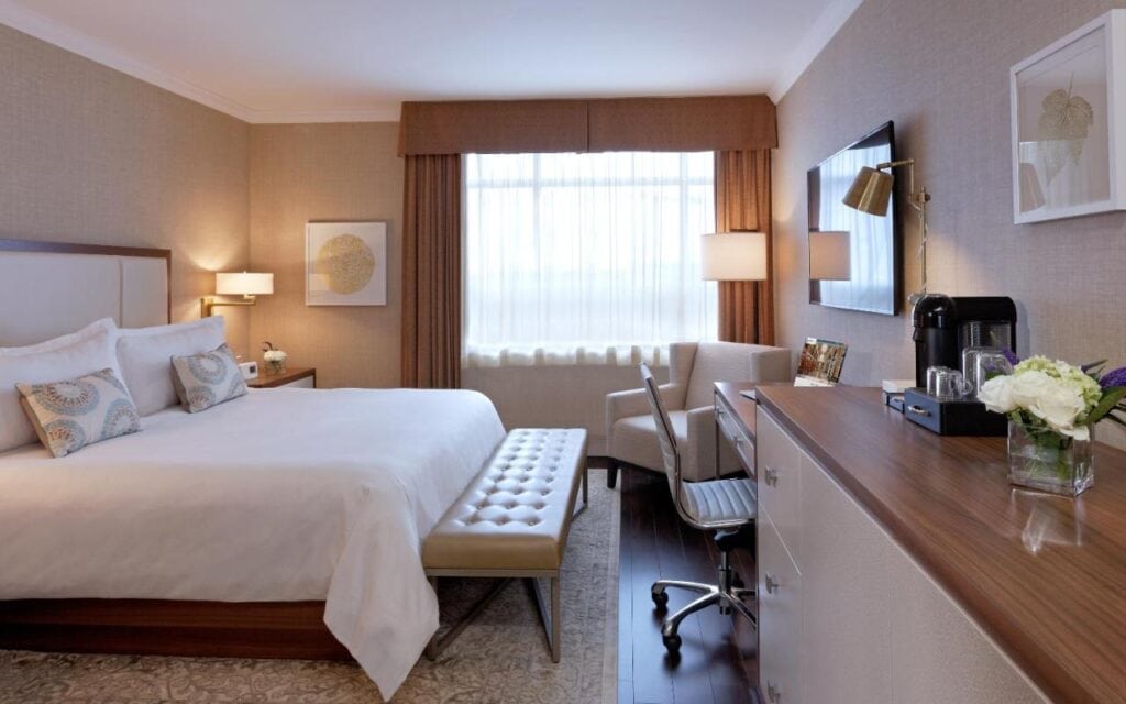 Where to Stay in Halifax: 17 Best Hotels in 2024 4 A large bed with white sheets and pillows inside a room. An ottoman is placed at the bed's end. A window with brown curtains, a single chair, and a desk and a swivel chair could also be seen. This is in Lord Nelson Hotel, one of the best places to stay in halifax.