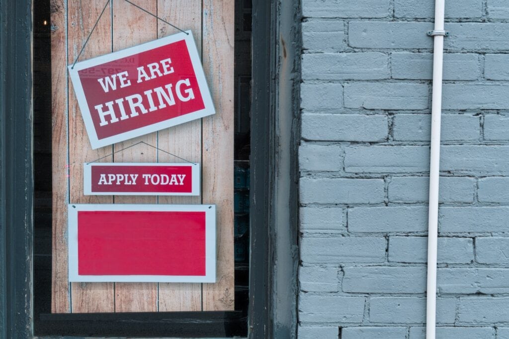 23 Top Pros and Cons of Living in Miami in 2024 7 A sign that says "we are hiring" and "apply today" hanging on a wooden door beside a brick wall. Miami is a city known to have a stable job market which is definitely a pro!
