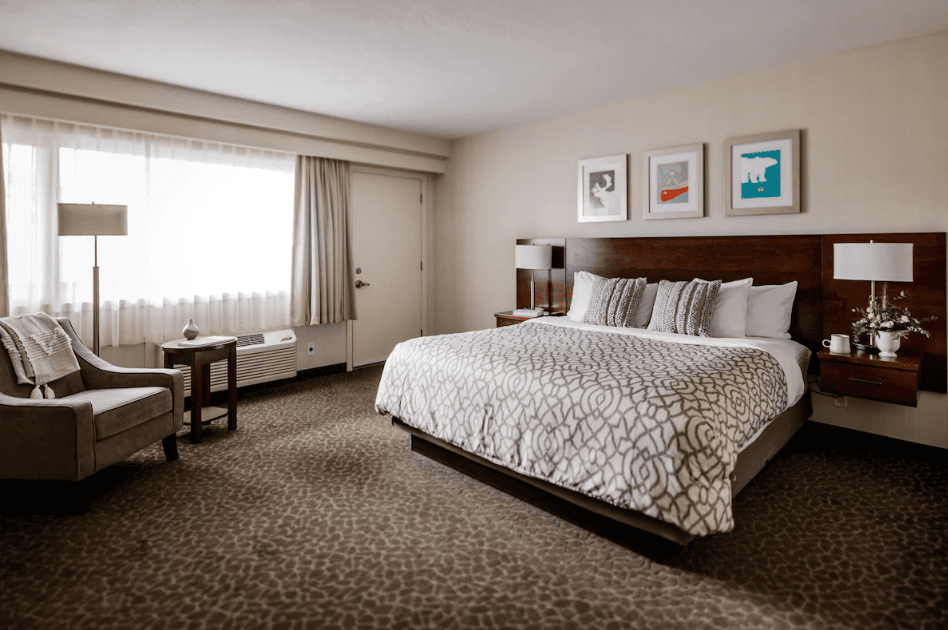 Where to Stay Near Jasper National Park: 16 Best Hotels 9 A large bed with patterned sheets in the middle od a room with white walls and carpeted floor. A single chair can be seen by the end of the bed near a wide window. This is one of the rooms of The Crimson Jasper in Jasper national Park
