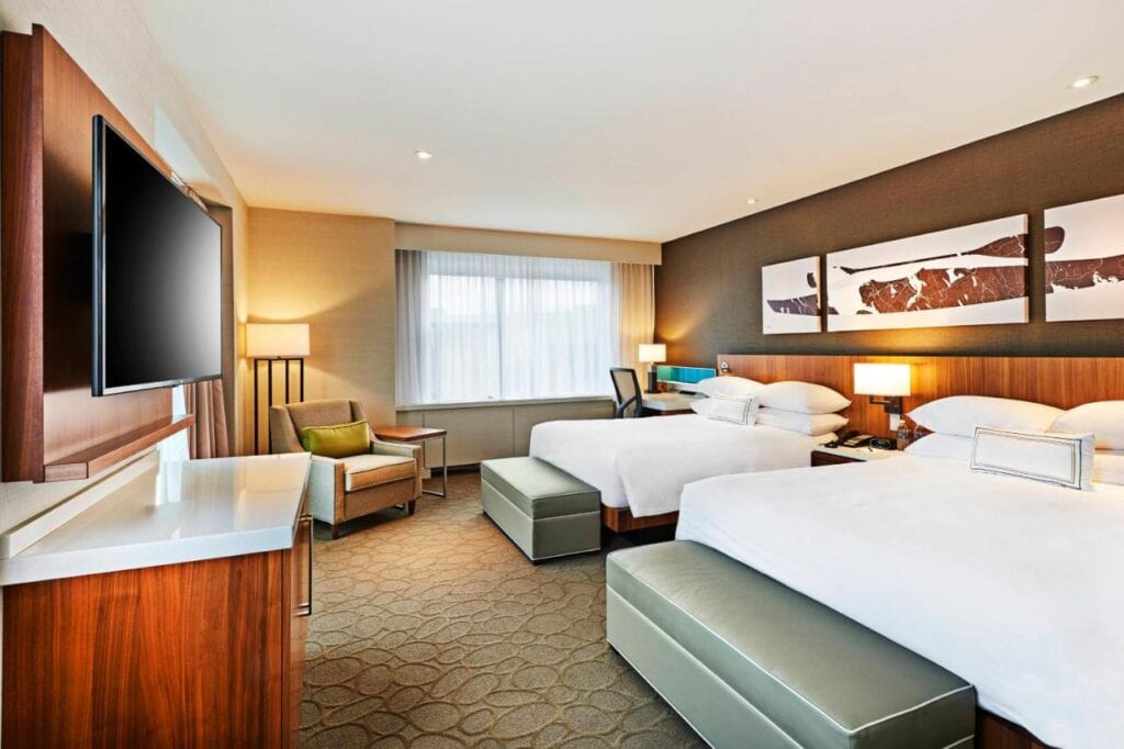 Where to Stay in Halifax: 17 Best Hotels in 2024 16 A photo of two large beds with white sheets in the middle of a well-lit room with brown walls. An ottoman is also placed at the end of each bed and a TV is mounted on a wall, facing the bed. This is taken from one of the rooms in Delta Hotels, one of the best places to stay in Dartmouth Halifax.