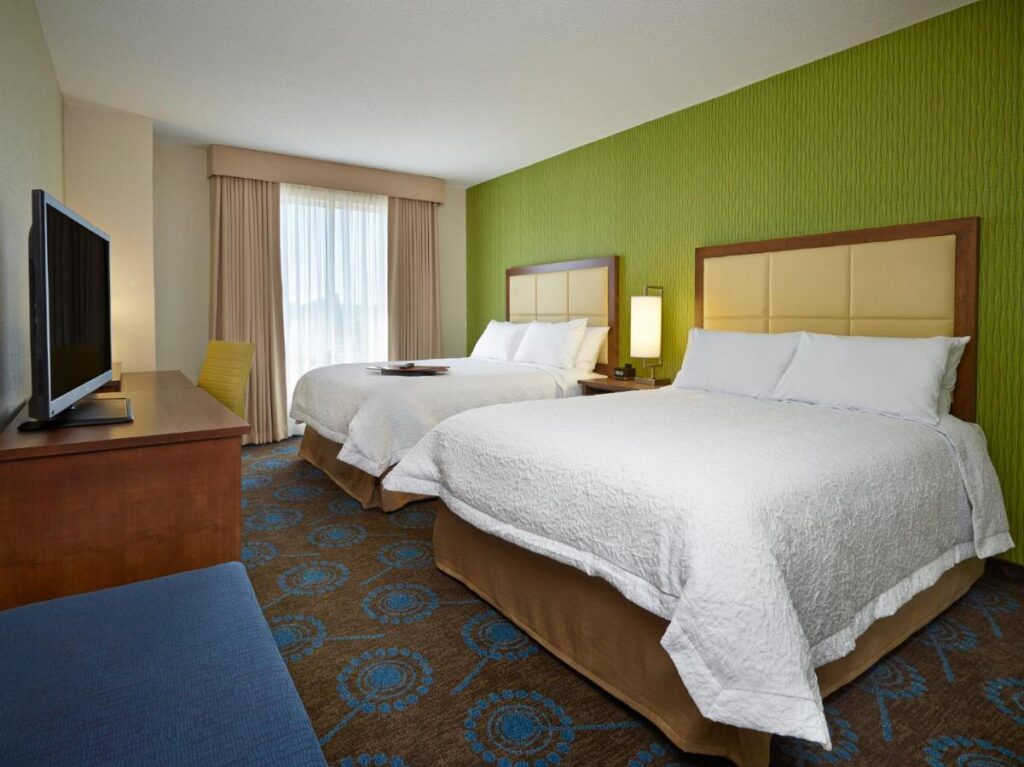 Where to Stay in Halifax: 17 Best Hotels in 2024 7 Two large bed with white matresses inside a room with green and beige walls. The floor is carpeted with a blue and brown design. It also has a TV ad a large window. This is the Hampton Inn, one of the best places to stay in Halifax.