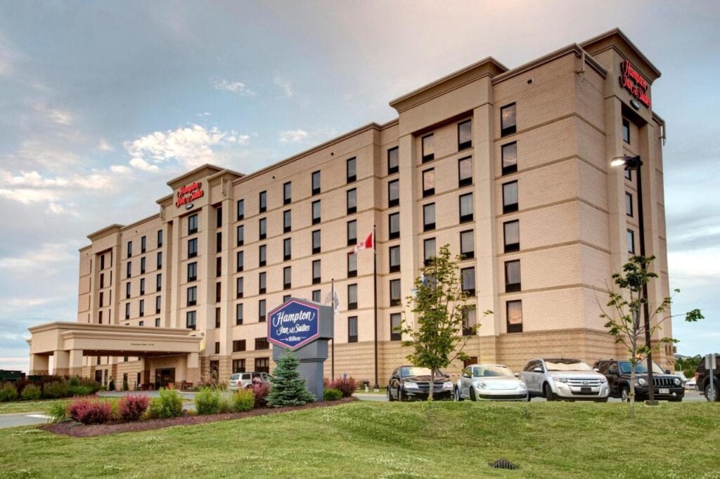 Where to Stay in Halifax: 17 Best Hotels in 2024 15 A photo of the outside of a beige building with windows lining up on its many floors. Several cars are also parked on the parking lot neer the grassy fields with a sign that says "Hampton Inn and Suites" This is one of the best places to stay in Halifax.