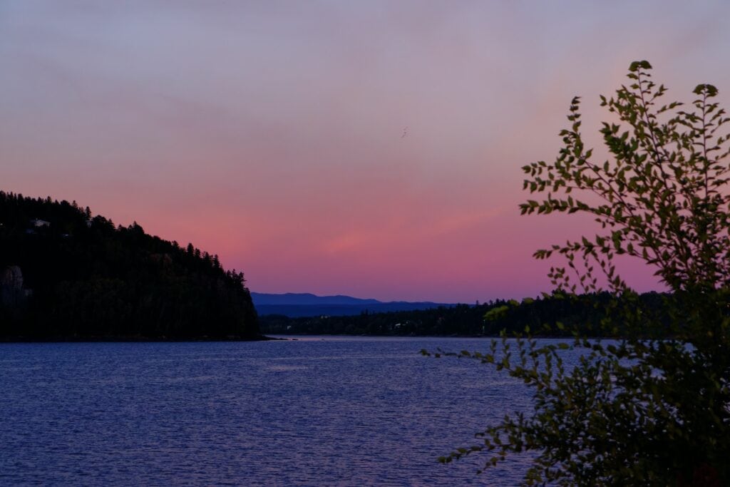 Whale Watching in Quebec: 9 Best Tours in 2024 12 A large body of water under a purple and pink sunset sky. A massive portion of land filled with trees is surrounding it. This is a photo of Fjord du Saguenay, a great place to go whale watching in Quebec.