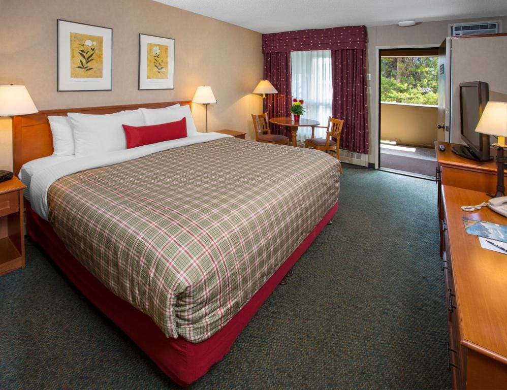 Where to Stay Near Jasper National Park: 16 Best Hotels 3 A large bed wth brown plaid sheets placed in the middle of a room with beige walls an a gray carpeted floor. Bedside tables with amps can also be seen on both sides of the bed near the curtained window. A balcony is also in selected rooms of the Marmot Lodge in Jasper.
