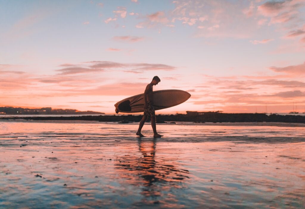 25 Pros and Cons of Living in Costa Rica in 2024 6 Man walking on the surf at sunrise with pale pink sky as he carries a surfboard.