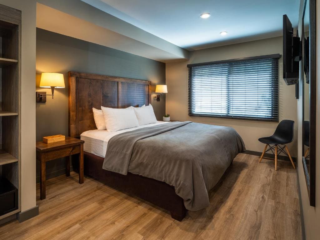 Where to Stay in Banff National Park: 18 Best Hotels (2024) 13 A large bed with white and grey sheets inside a room with gray walls and wooden floor. A window with blinds can be seen beside the bed and near a black chair. A TV is mounted on a wall, facing the bed. This is in Buffalo Mountain Lodge, a hotel near Banff national Park.