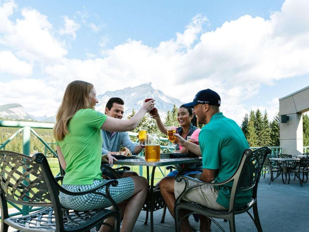 Where to Stay in Banff National Park: 18 Best Hotels (2024) 1 A group of people sitting on chairs surrounding a round table, drining beer. A view of a large mountain can be seen in the background. This is in the Banff Springs Hotel, one of the best hotels in Banff National Park.