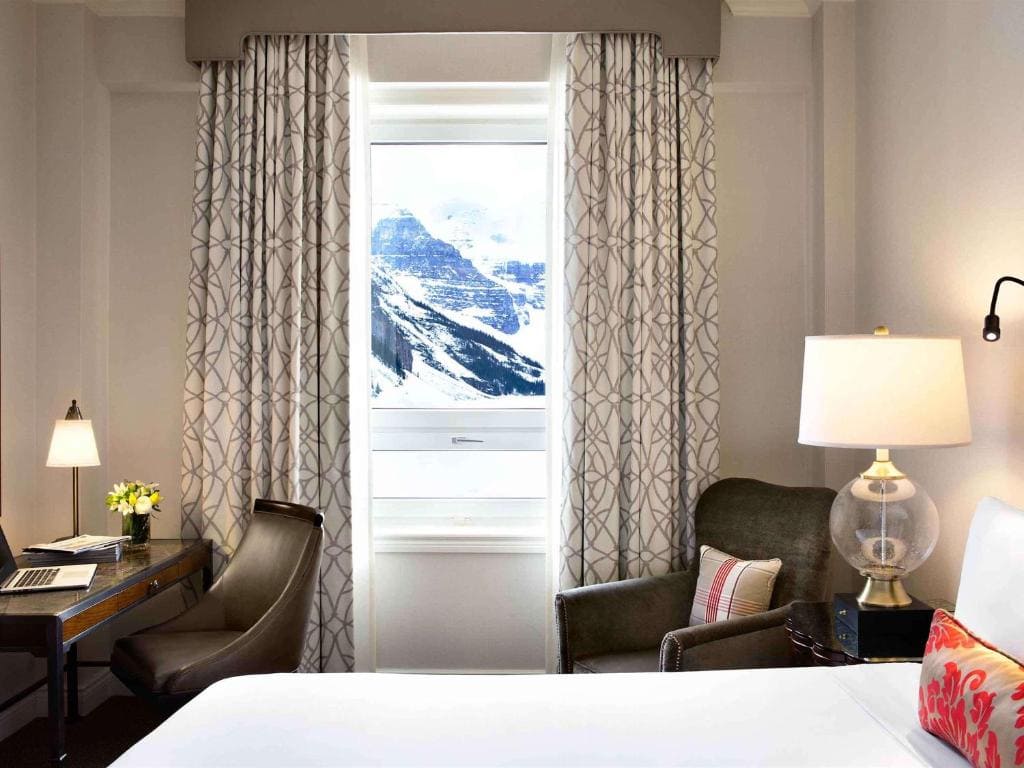 Where to Stay in Banff National Park: 18 Best Hotels (2024) 17 A beige-colored room with a bed, a table, and two chairs beside a window with patterned curtains. A snow-covered mountain can be seen from the window. This is the view from one of Fairmont Chateau Lake Louise's rooms.