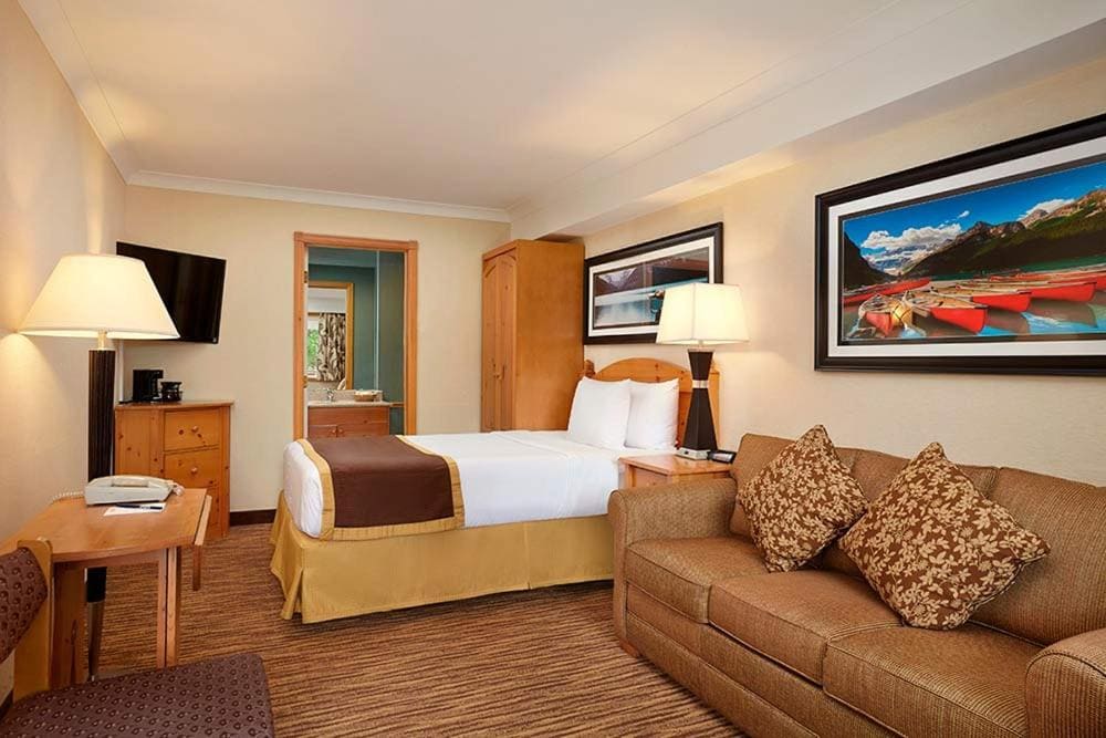 Where to Stay in Banff National Park: 18 Best Hotels (2024) 18 A photo of a room wth beige walls and a carpeted floor. A bed can be seen in the middle of the room beside a brown couch, a cabinet, and a bedside table with a lamp. This is in Lake Louise Inn, one of the best places to stay near Banff National Park.