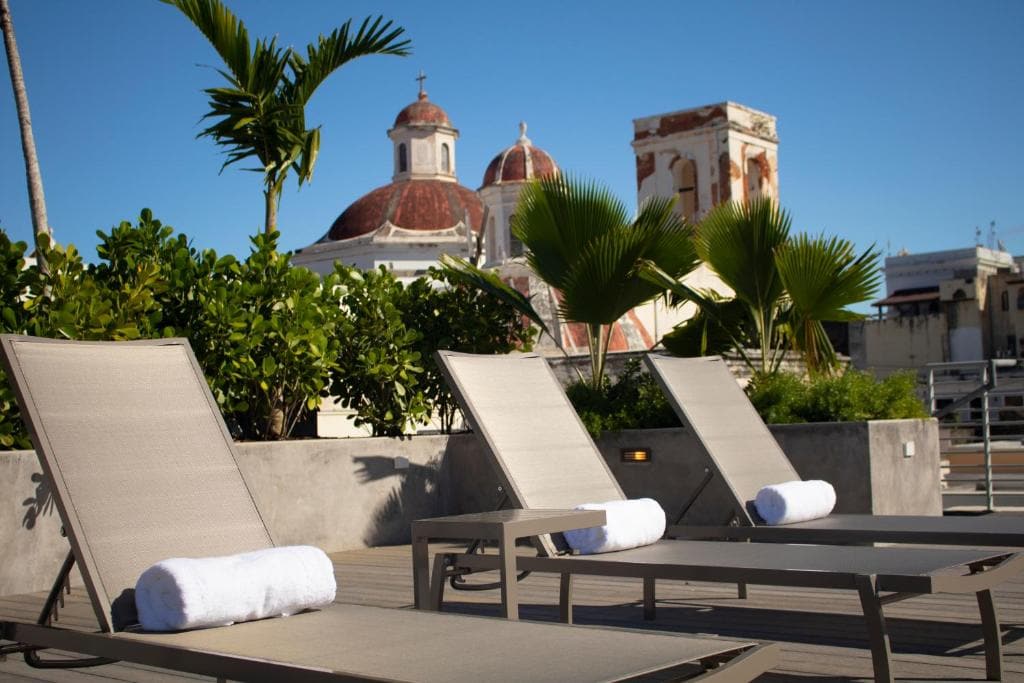 13 Best Hotels in Old San Juan, Puerto Rico 2024 7 Three bathing chars with towels lining up near a large pot of green plants. The dome-shaped roof of a church can also be seen in the background This is in Placio Provincial, a luxury hotel in San Juan, Puerto Rico.