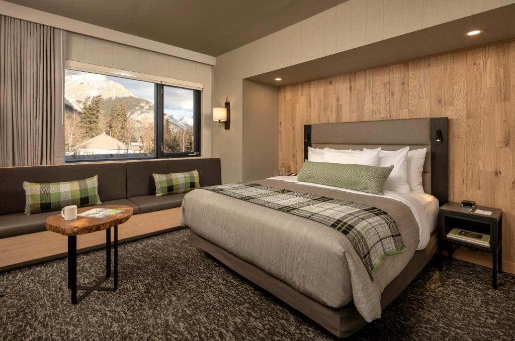 Where to Stay in Banff National Park: 18 Best Hotels (2024) 9 A bed with grey and green sheets inside a brown colored room and a carpeted floor. An upholstered couch can be seen beside the bed and below the window with the view of the majestic nature. This is in The Peaks Hotel and Suites in Banff.