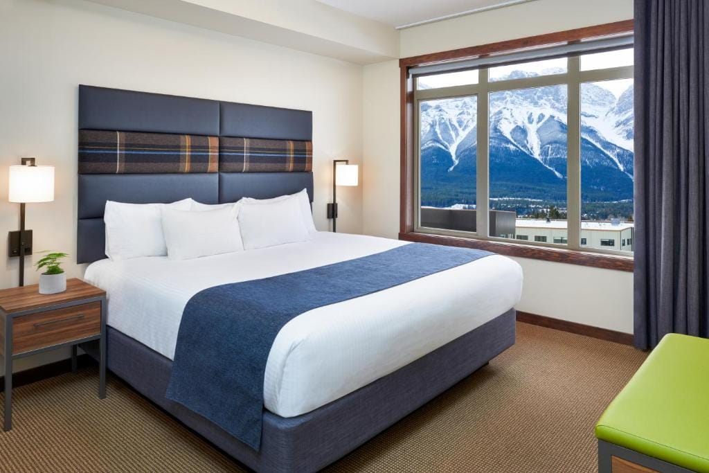 Where to Stay in Banff National Park: 18 Best Hotels (2024) 21 A room with white walls and brown carpeted floor with a large bed in the middle. A huge window with a view of a snow covered mountain can be seen beside the bed. This is in Stoneridge Mountain Resort.