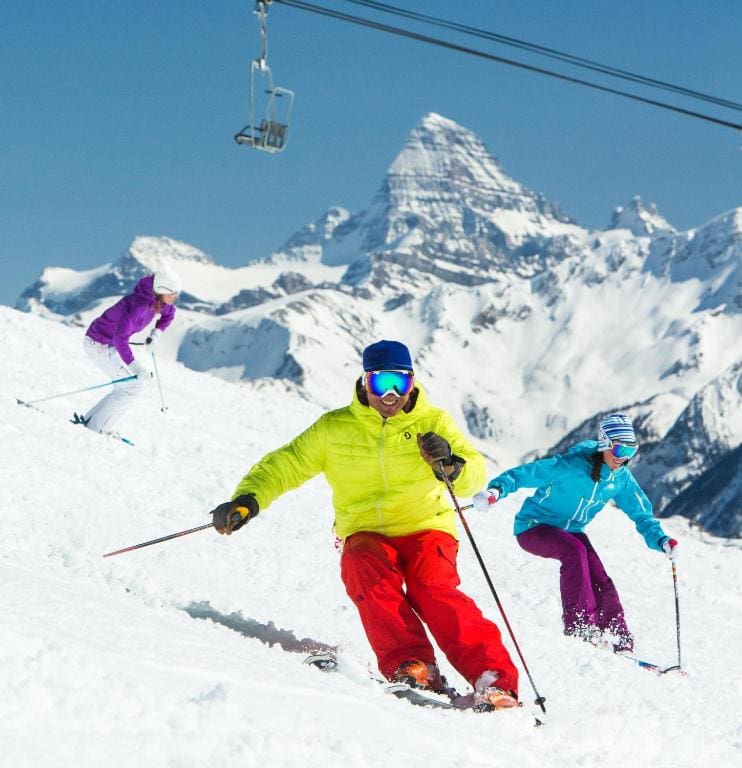 Where to Stay in Banff National Park: 18 Best Hotels (2024) 16 Three people wearing snowgear and skiing equipment skiing down a snow-covered mountain. This is one of the activities near Sunshine Mountain Lodge, a hotel near Banff National Park.