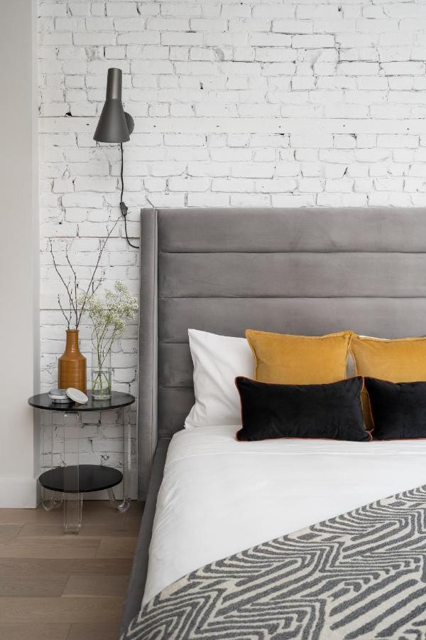 22 Best Places to Stay in Montreal Canada 13 A bed with white and grey sheets inside a room with white brick walls and vinyl tiles flooring. A bedside table with vases of flowers can be seen beside the bed. This is the Francois-Denis Apartments in Montreal Canada.