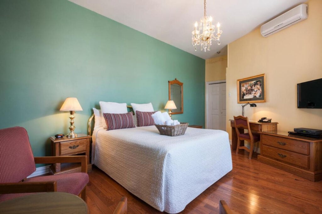 22 Best Places to Stay in Montreal Canada 22 A room with pastel colored walls and wooden floors that has large bed inside. A TV can be seen on top of the dresser near the bed, as well as a desk and a chair. This comfortable room is in the Hotel Chateau de L'Argoat in Montreal Canada.