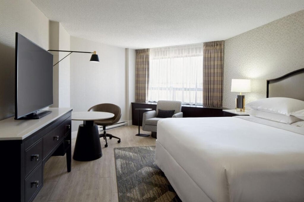 22 Best Places to Stay in Montreal Canada 25 A room with white walls and wooden flooring with a large bed inside. In front od the bed is a TV Stand that doubles as a dresser, a flat screen TV, a round desk, and a swivel chair. This is in Sheraton Montreal airport in Montreal, Canada.