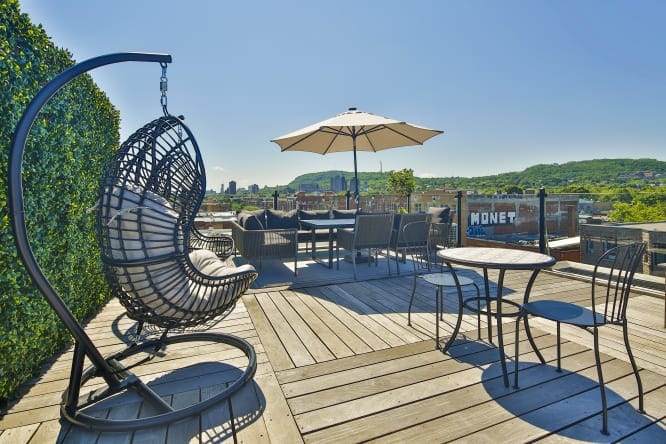 22 Best Places to Stay in Montreal Canada 16 Outdoor chairs and tables with umbrella in a deck with wooden flooring. A view of the city and the green trees can be seen from the deck. This is in the Parc Avenue Hostel in Montreal Canada