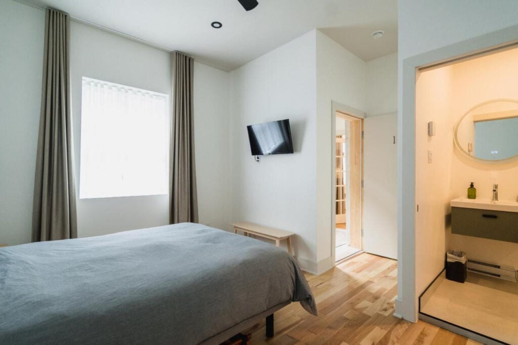 22 Best Places to Stay in Montreal Canada 18 A room with white walls and laminated flooring with a bed with gray sheets inside. A flat screen wall-mounted TV can be seen in front of the bed.