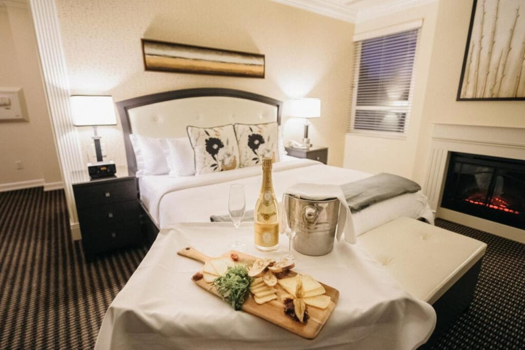22 Best Places to Stay in Montreal Canada 3 A large bed inside a carpeted floor with white floors and two bedside tables and lamps in both sides. A serving cart of a charcuterie board, champagne, and ice bucket can also be seen in the photo. This is one of the best places to stay in Montreal Canada.