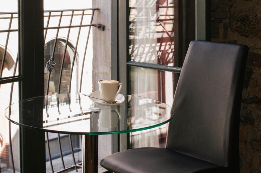 22 Best Places to Stay in Montreal Canada 8 A round glass table with a coffee cup and saucer and a black leather chair near a sliding window. This is in Le Petit hotel, one of the best places to stay in Montreal Canada.