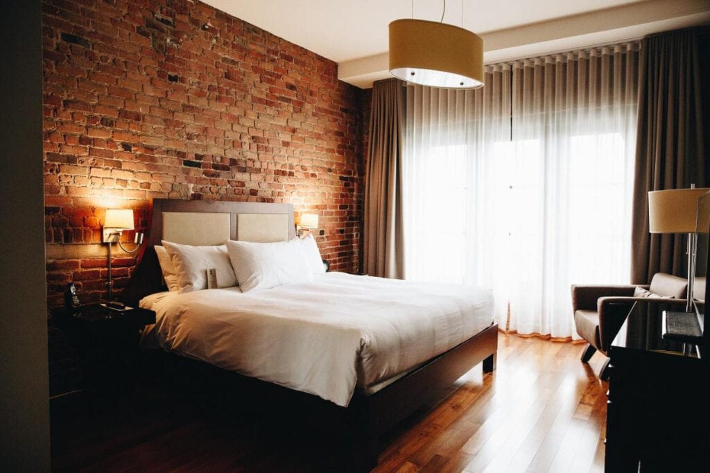 22 Best Places to Stay in Montreal Canada 9 A large bed with white sheets inside a room with laminated wooden floors and brick walls. A flat screen TV is placed in front of the bed and a large window with curtains on the left side of the room. This is the luxurious Hotel Nelligan in Montereal Canada.
