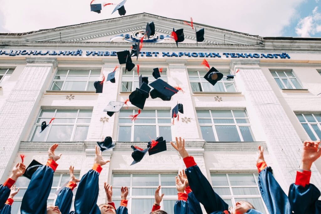 25 Pros and Cons of Living in New Hampshire in 2024 9 Students in black and orange togas throwing their graduation caps in the air in fromnt of a large white building. One of the good things about living in New Hampshire is its quality education for the learners.
