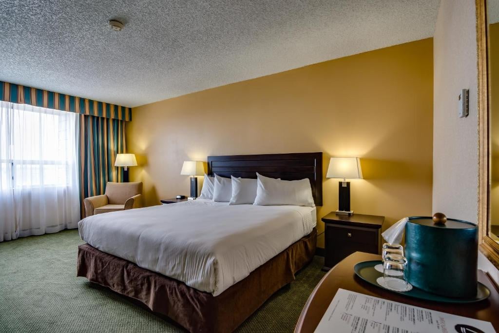 11 Best Edmonton Hotels With Balconies 2024 6 A large bed with four white pillows inside a carpeted room. Two bedside tables and lamps are placed on both sides of the bed. This is in the Sawridge Inn in Edmonton.