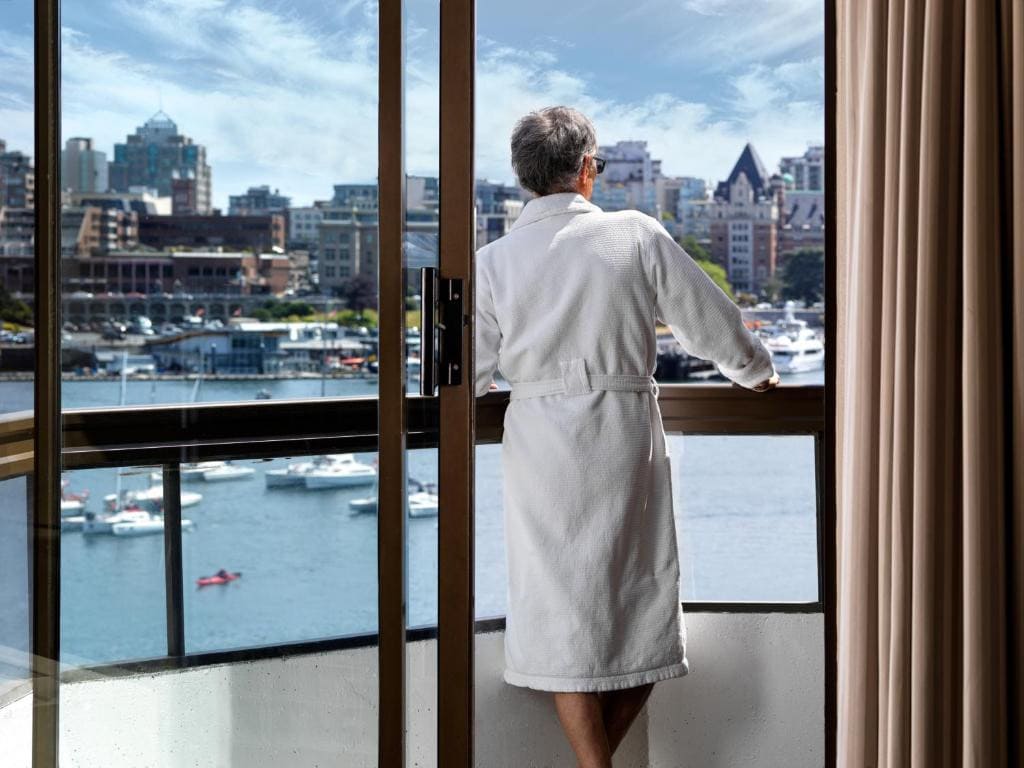 21 Best Places To Stay In Victoria, Canada 2024 10 A man in white bathrobe looking at the balcony view of a large body of water near a city with tall buildings. The Inn at Laurel Point is one of the best places to stay in Victoria, Canada.