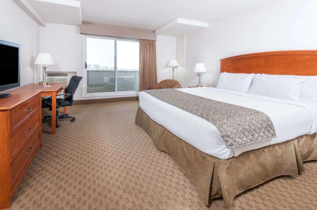 11 Best Edmonton Hotels With Balconies 2024 8 A large bed with white sheets facing a television placed above a wooden dresser. Tall lamps can be seen in the corners of the room and a balcony is on the right side of the bed. This is in the Ramada by Wyndham in Edmonton.