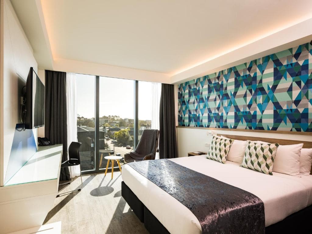 18 Best Hotels in Christchurch, New Zealand 2024 5 A bed facing a dresser and a wall-mounted TV unsde a room with wooden flooring. Beside the bed is a table, a chair, and a large window offering a stunning view. This is Carnmore hotel, one of the best places to stay in Christchurch