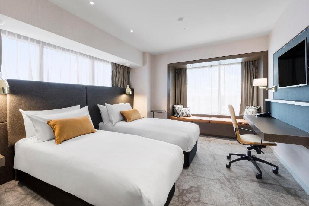 18 Best Hotels in Christchurch, New Zealand 2024 7 Two beds facing a swivel chair and a wall mounted TV inside a carpeted room. A large window can be seen beside one bed, This is in the Crowne Plaza in Christchurch.