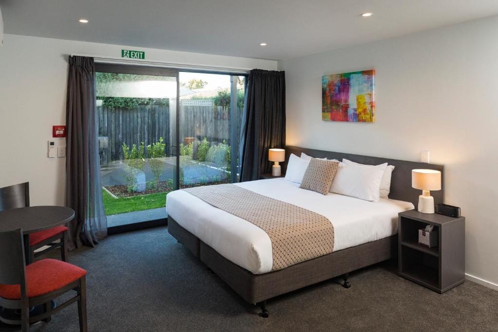 18 Best Hotels in Christchurch, New Zealand 2024 13 A table and two chairs in front of a bed inside a carpeted room. Beside the bed is a sliding door towards a yard. This is in the Carnmore hagley park in Christchurch, New Zealand.