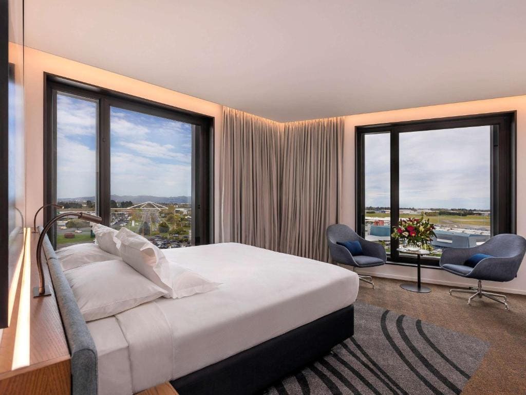 18 Best Hotels in Christchurch, New Zealand 2024 17 A bed facing a round table with two gray chairs. Large windows can be seen, offering a stunning view. This is in the novotel Christchurch Airport Hotel.