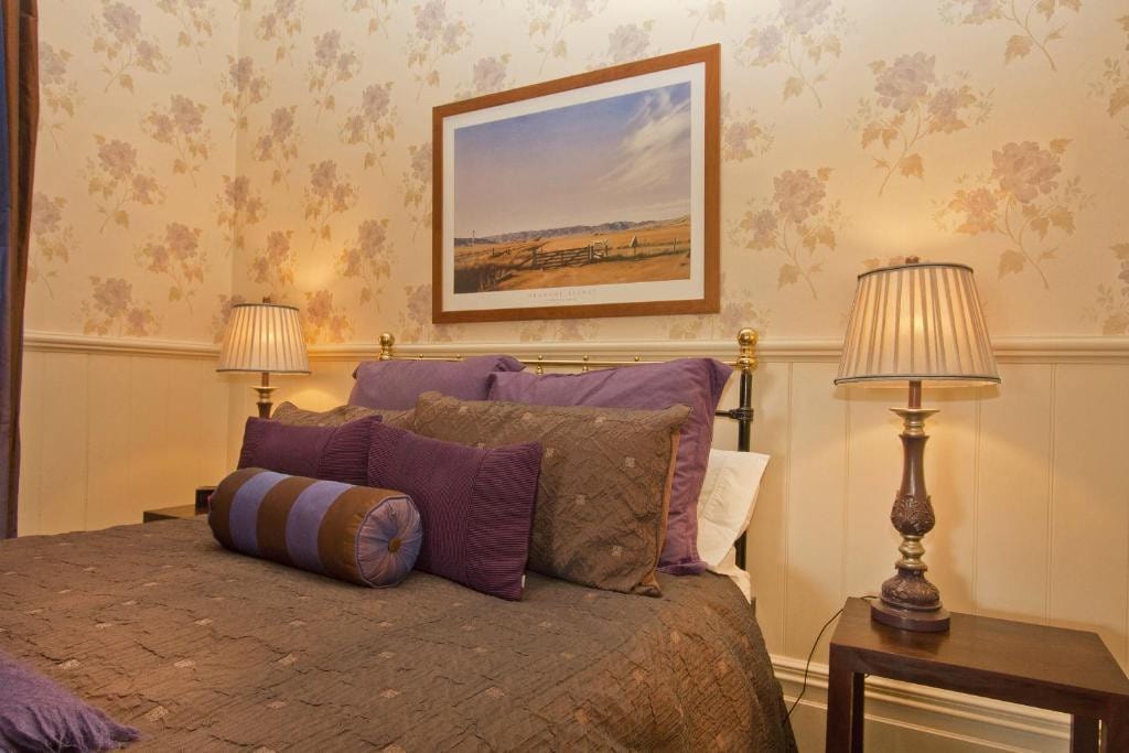 18 Best Hotels in Christchurch, New Zealand 2024 18 a bed with brown and purple beddings inside a room with wallpaper and a picture frame above the bed. This is in Lilac Rose Boutique Bed and Breakfast in Christchurch.