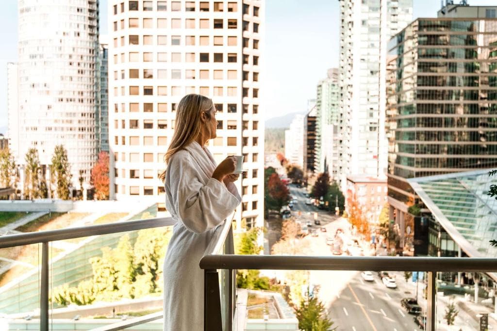 A woman wearing awhite robe looking over large buildings from a balcony with glass and metal railings. This is in SHangi-la vancouver, one of the best vancouver hotels with balconies.