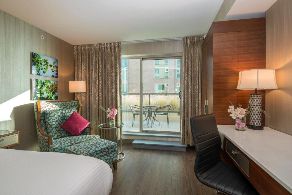 23 Best Vancouver Hotels with Balconies (A Local’s Guide) 8 A bed, a printed chai, a swivel chair, and a desk inside a room with wooden flooring. A sliding door towards a balcony can be seen near the chair.