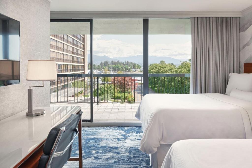 23 Best Vancouver Hotels with Balconies (A Local’s Guide) 14 Two beds inside a carpeted room in front of a desk and a wall-mointed TV. A sliding door towards a balcony with metal railings can be seen beside one bed. This is in Westin Bayshore, Vancouver.