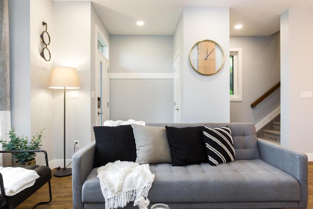 11 Best Edmonton Hotels With Balconies 2024 13 A gray couch with black and gray couch pillows on a living room. A tall lamp can be seen behind the couch and a clock is mounted on a wall near the stairs on the right side. This is the Contemporary Farmhouse in Edmonton.