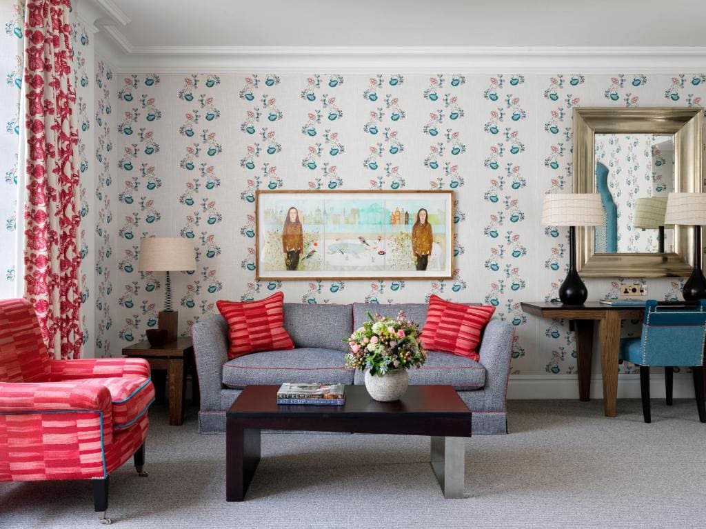 27 Best London Hotels with Balconies 2024 4 A living room with floral wallpaper and carpeted floor. There is a gray couch and a red solo couch, and a black coffee table with a vase on the center. This is in the Soho Hotel one of the best london hotels with balconies.