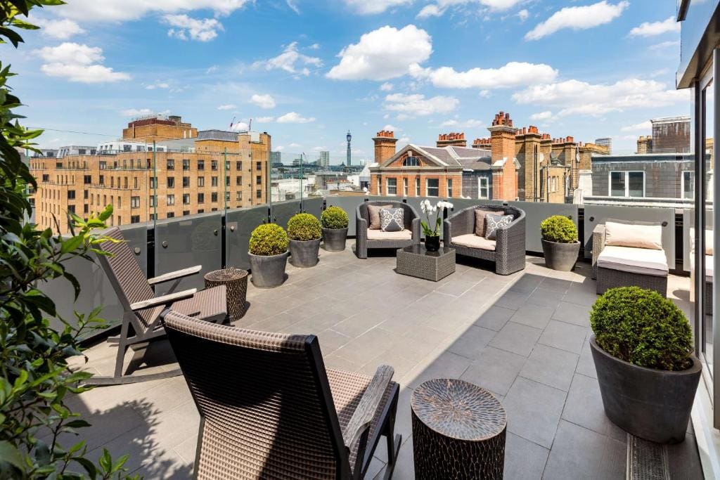 27 Best London Hotels with Balconies 2024 6 A balcony with gray tiled flooring and cement railings with a view of large buildings. Several outdoor rattan chairs and outdoor plants can be seen in the photo. This is the May Fair in Westminster Borough, London.
