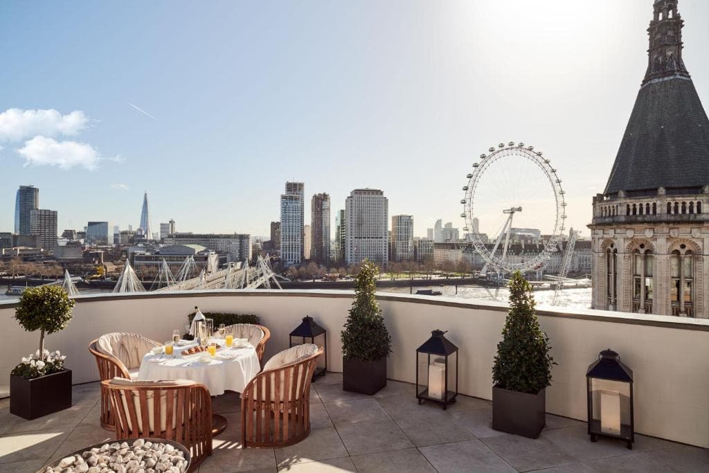 27 Best London Hotels with Balconies 2024 7 A balcony with gray tales and concrete railing with a view of large buildings, a bridge, and a ferris wheel. A table with wooden chairs are also set up in the balcony. This is in the COrinthia London.