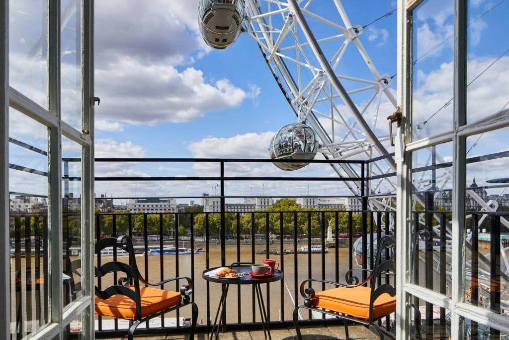 27 Best London Hotels with Balconies 2024 9 A balcony with wooden flooring and black metal railings with a view of a large body of water and a ferris wheel. Two orange chairs and a a small round table can be seen. This is in the London Marriott Hotel County Hall, London.