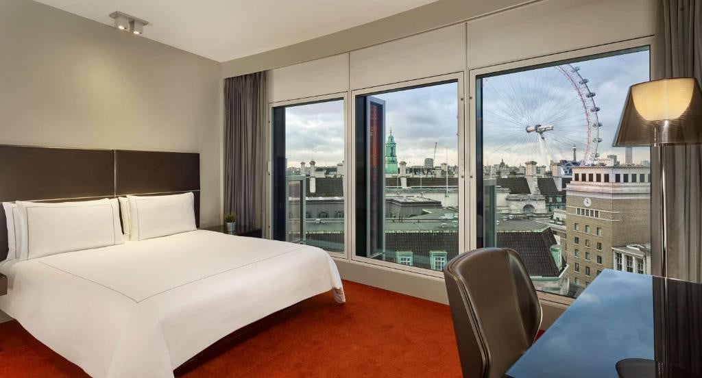27 Best London Hotels with Balconies 2024 11 A hotel room with carpeted floors and white walls, with a large bed beside huge windows, offering a view of buildings and a ferris wheel. This is in the Park Plaza Westminster Bridge, London