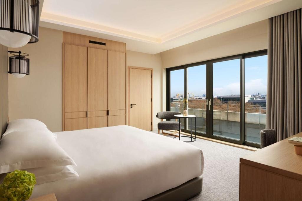 27 Best London Hotels with Balconies 2024 15 A hotel room with a large bed beside wooden cabinets and a dresser. In front of the bed, is a sliding door to a balcony overlooking a view of the city, this is the Nobu Hotel, one of the best london hotels with balconies.