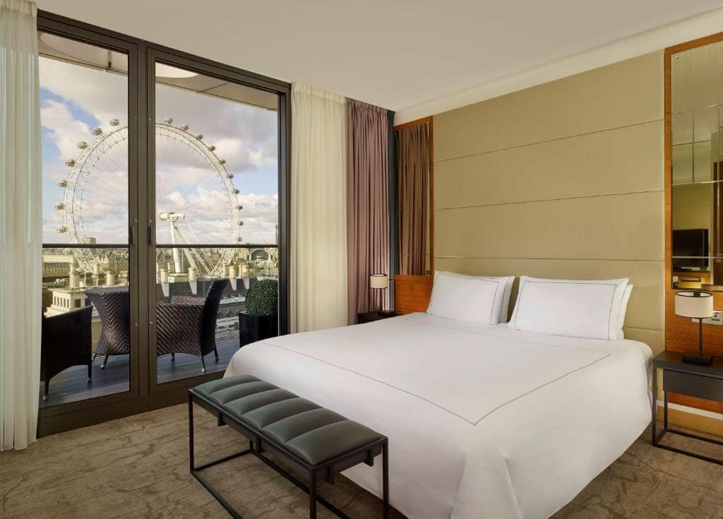 27 Best London Hotels with Balconies 2024 16 A hotel room with a large bed with white sheets beside a glass door to a balcony with rattan chairs and tables, overlooking a view of large buuldings and a ferris wheel. This is in the Park Plaza County Hall London.