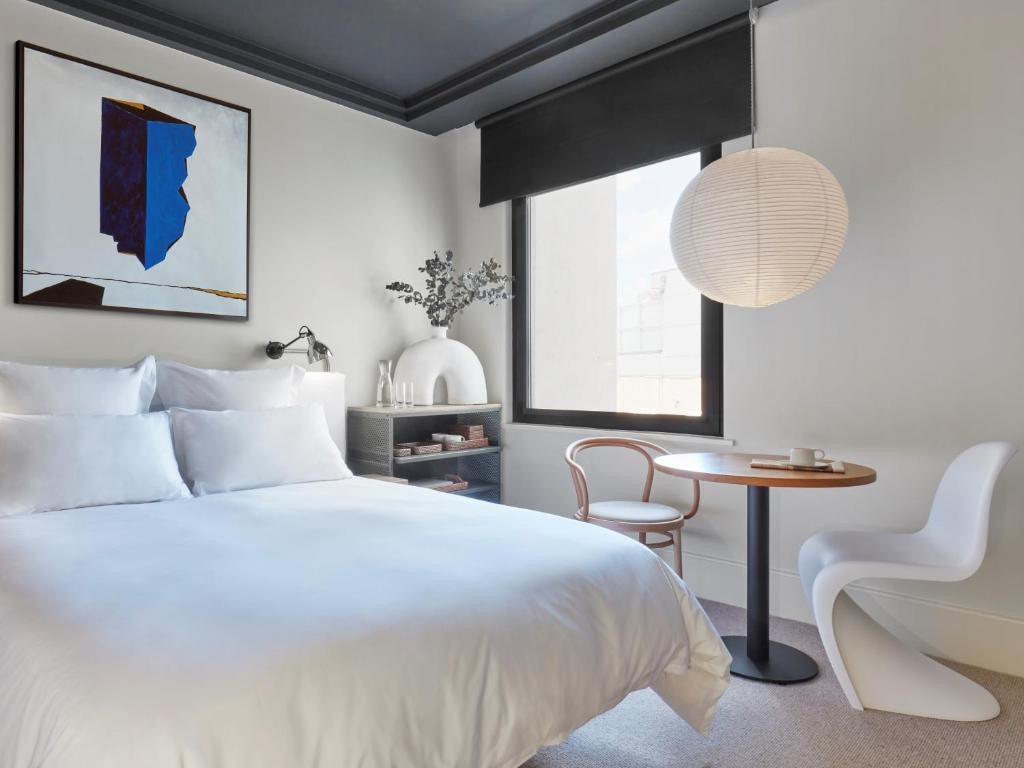 27 Best London Hotels with Balconies 2024 17 A white bed inside a room with white walls and beside a round table with two different types of chairs. Near the table is a square window. This is in the One Hundred Shoreditch, London.