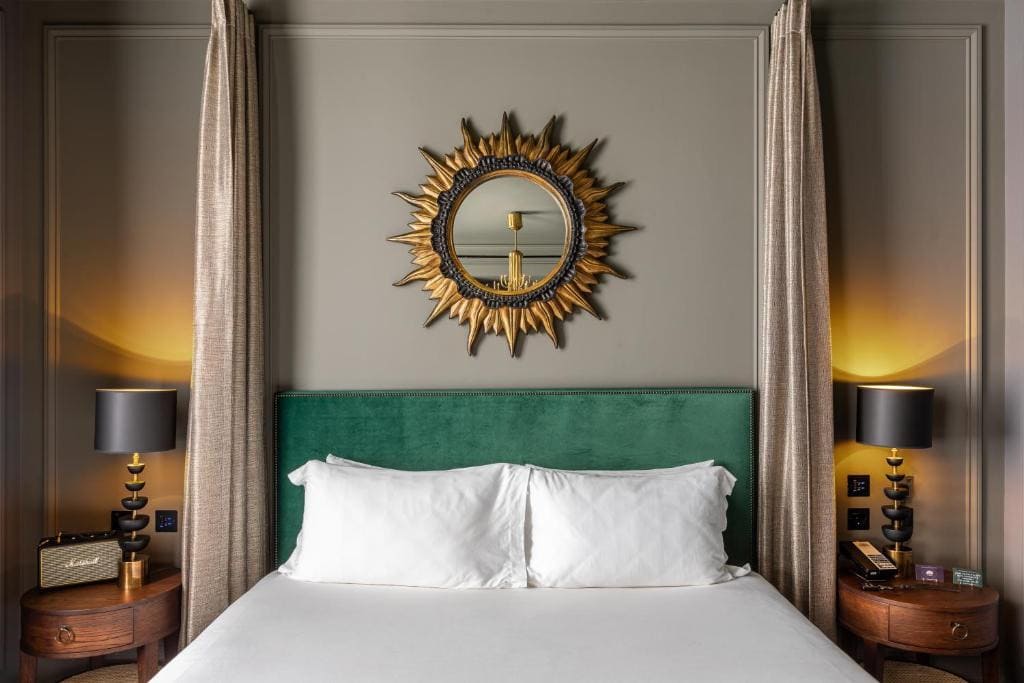 27 Best London Hotels with Balconies 2024 19 A bed in a room with a green headboard and a round sun-shaped mirror, and two bedside tables on both sides of the bed. This is in The Mandrake, London.